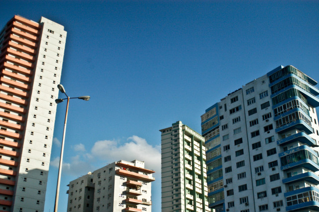 A group of colorful high-rise residential buildings, featuring distinct architectural designs, stands against a clear blue sky. The scene includes a streetlight in the foreground.