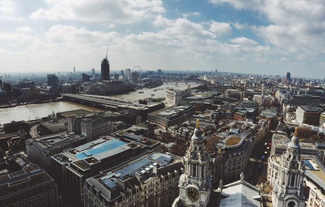 Aerial view of central London, showcasing iconic landmarks like the River Thames, London Eye, and St. Paul's Cathedral. Urban architecture, bridges, and bustling city life are visible under a partly cloudy sky.