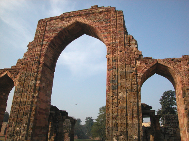 A close-up view of a massive, red sandstone archway in Delhi, India. The archway is adorned with intricate carvings and designs, showcasing the architectural beauty of the Qutub Minar complex.