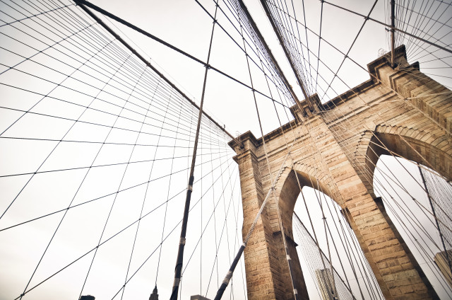 Low-angle view of the Brooklyn Bridge in New York City, featuring its stone arches and converging suspension cables. The geometric lines of the bridge create a symmetrical pattern against the bright sky.