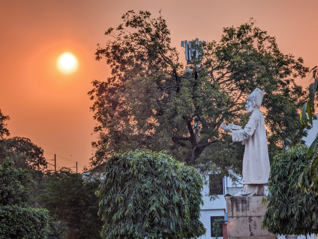 The statue of renowned poet Ghalib stands in the gardens of Jamia Millia Islamia as the sun sets, casting an orange glow across the sky. The peaceful scene is framed by lush greenery and tall trees, enhancing the serene atmosphere.