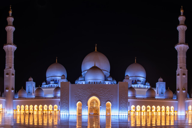 A breathtaking nighttime view of the Sheikh Zayed Grand Mosque in Abu Dhabi, UAE. The mosque is illuminated with white light, highlighting its intricate architectural details and towering minarets