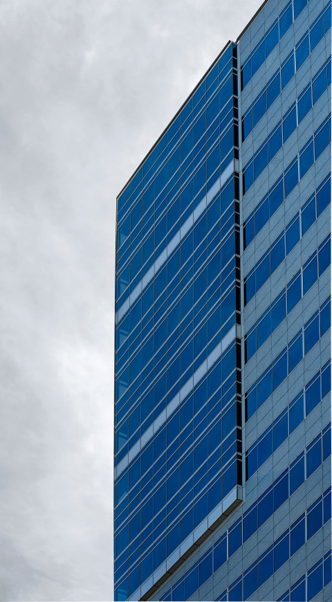 A close-up of a modern corporate building featuring a blue glass facade with horizontal lines and a cloudy sky in the background