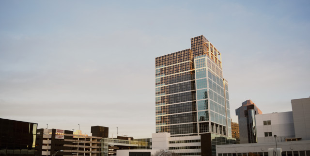 A modern glass-clad office building reflects sunlight in an urban environment, accompanied by a multi-level parking structure labeled "Discount Parking." The scene captures a blend of architecture under a clear sky