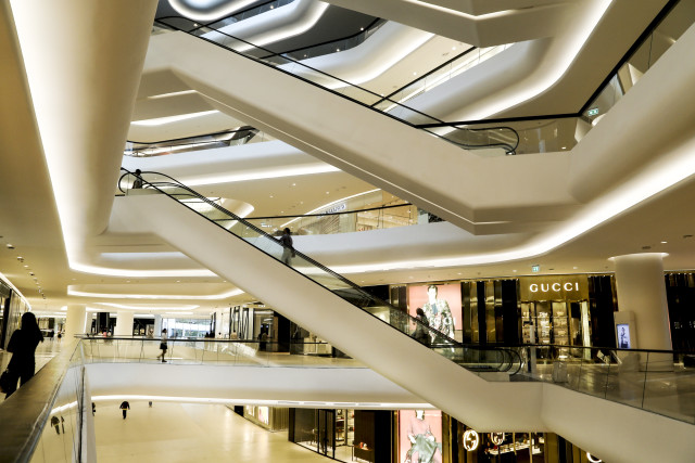 Luxurious multi-level shopping mall with sleek white architecture, illuminated interiors, escalators, and high-end stores including Gucci. Clean, contemporary design with open spaces and minimal foot traffic.