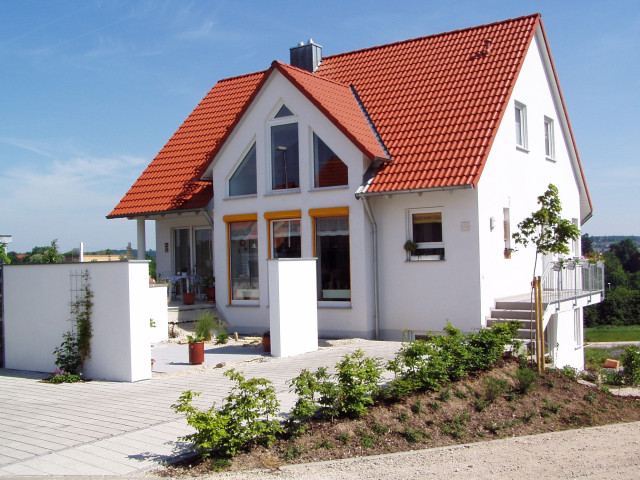 A modern white house with a vibrant red gable roof, large triangular windows, and a well-maintained garden featuring shrubs and a paved driveway. Located in a suburban area with clear skies in the background