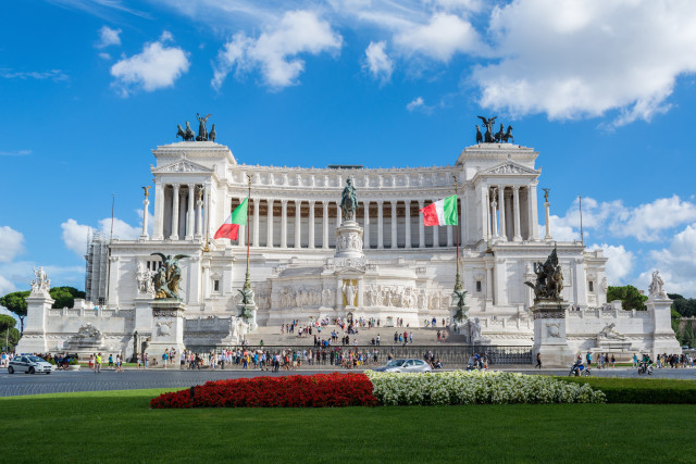 A grand white marble monument stands tall in Piazza Venezia, Rome, Italy. The monument is adorned with statues and sculptures, and the Italian flag flies proudly above. The monument is surrounded by lush green lawns and a bustling street.