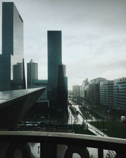 Urban scene of Rotterdam city center with tall skyscrapers, modern architecture, and a rainy street below. Wet roads and trams are visible alongside towering glass buildings under an overcast sky.