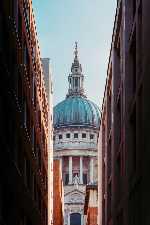 St. Paul’s Cathedral dome in London, framed by modern buildings on both sides, with the structure’s intricate architecture and classical detailing highlighted against a clear blue sky.