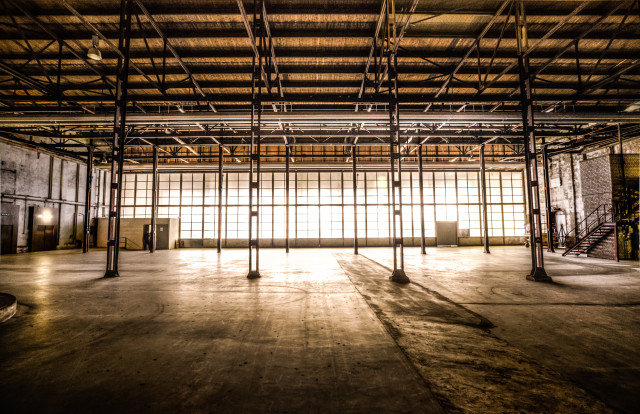 A large, empty industrial warehouse with high ceilings and a concrete floor. Sunlight streams through a row of windows, casting long shadows