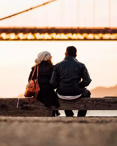 two people sitting on a bench looking at the bridge